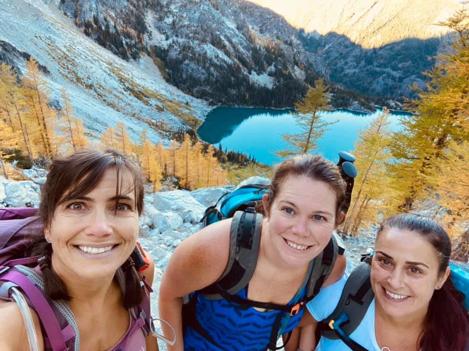 Three women with backpacks smile at the camera while hiking. Behind them, a mountainous landscape features a clear, blue lake surrounded by autumnal trees and rocky terrain under a bright sky.