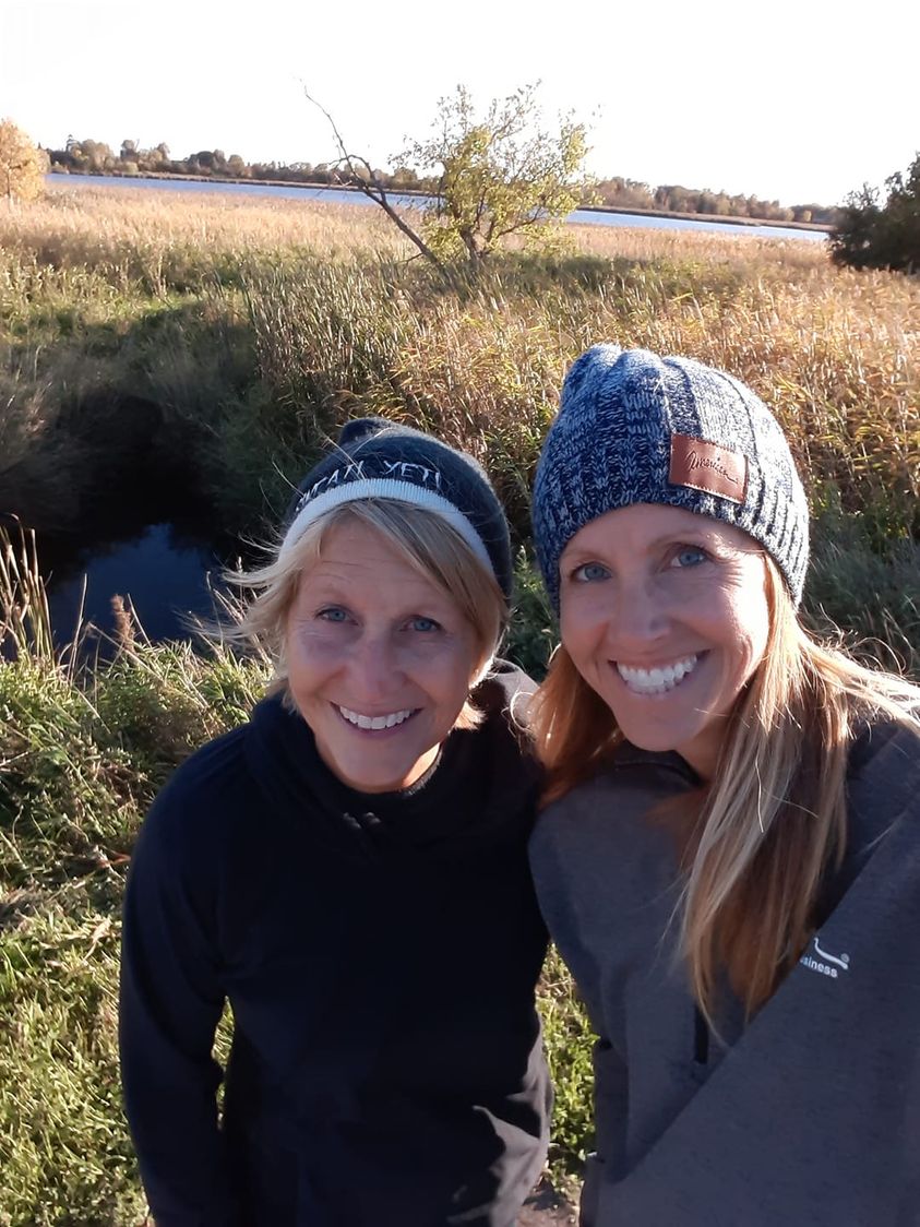 Two women wearing winter hats and jackets smile at the camera while standing outdoors in a grassy area. A small body of water and trees are visible in the background under a clear sky. The scene appears bright and sunny.