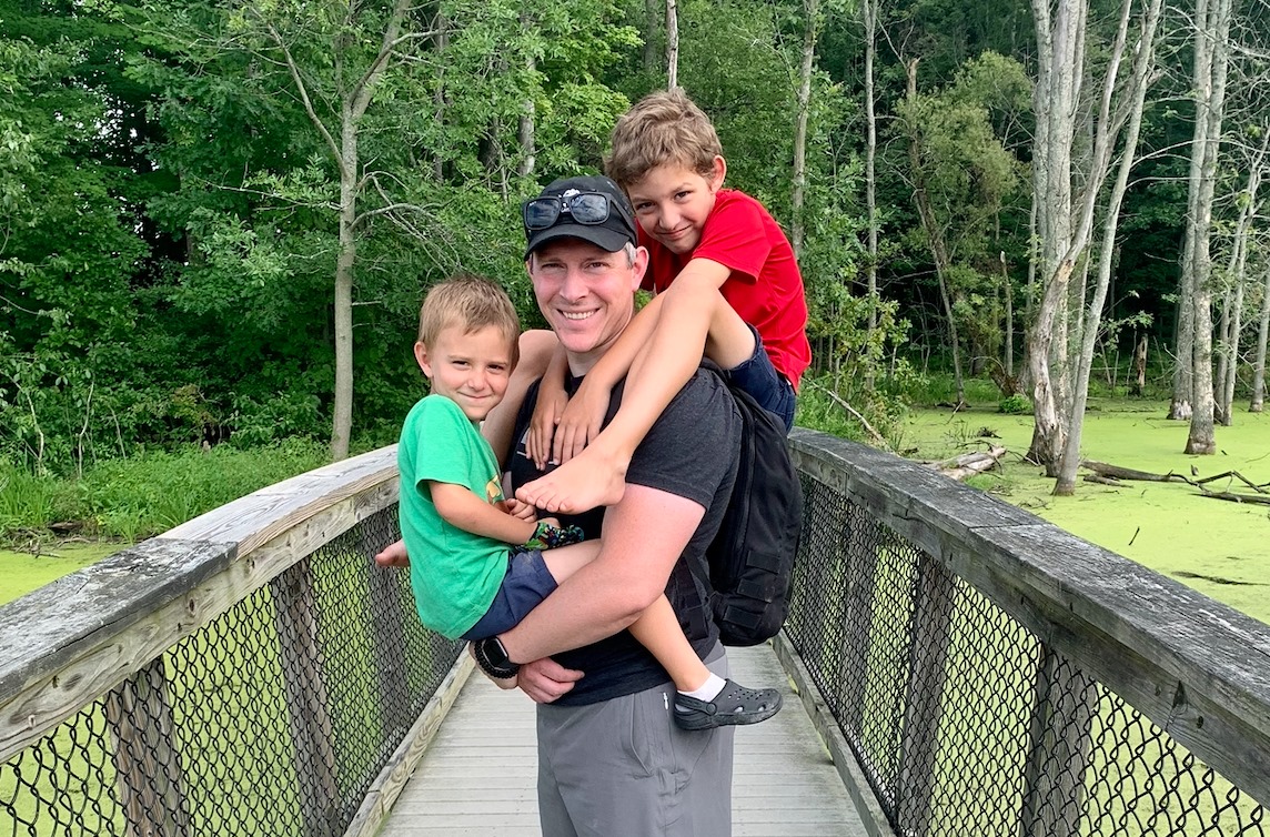 A man smiles while carrying two young boys on a wooden bridge. One boy, in a green shirt, is held in his left arm and the other boy, in a red shirt, is piggybacking. The bridge is surrounded by lush greenery and a waterbody covered in green algae.