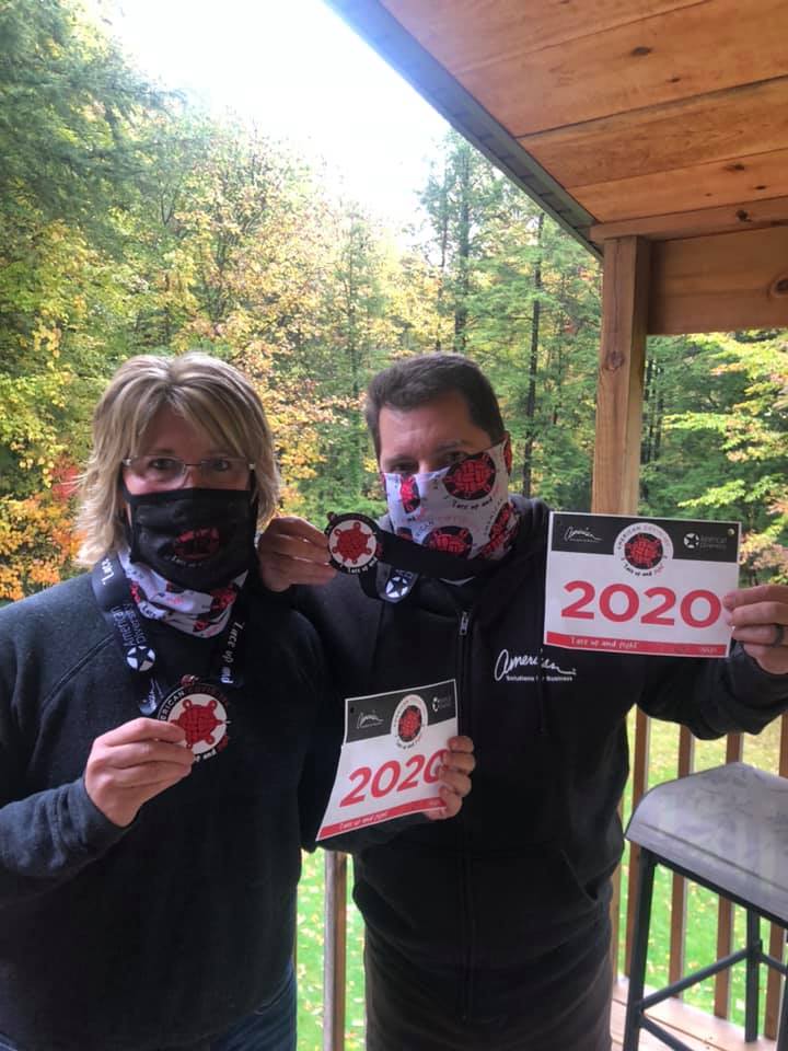 Two people stand on a porch wearing neck gaiters with a marathon logo. They both hold running bibs displaying "2020" and medals. They appear to be celebrating their participation in a race. The background features a wooded area with autumn foliage.