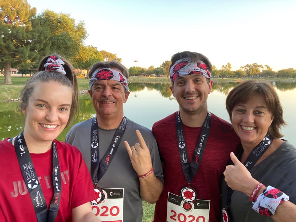 A group of four people, two men and two women, wear matching race bandanas and medals. They smile at the camera, standing near a lake with trees in the background. The medals read "2020." They are dressed in athletic gear, indicating participation in a race or event.