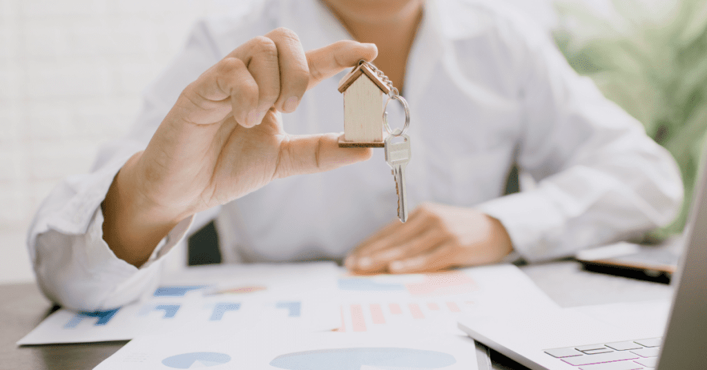 A person in a white shirt holds a house-shaped keychain with a key, sitting at a desk with graphs and charts, illustrating a unique story of real estate, home buying, or financial planning.