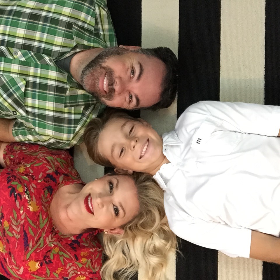 Three people are lying on a black and white striped surface. A man in a green plaid shirt, a woman in a red floral top, and a child in a white shirt are smiling at the camera. Their heads are close together, forming a triangle.