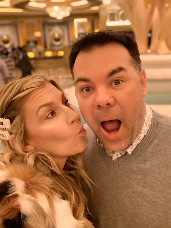 A man and woman take a selfie indoors. The woman with long blonde hair adorned with a hairclip is playfully leaning in to kiss the surprised-looking man on the cheek. The background features elegant decor and lighting.