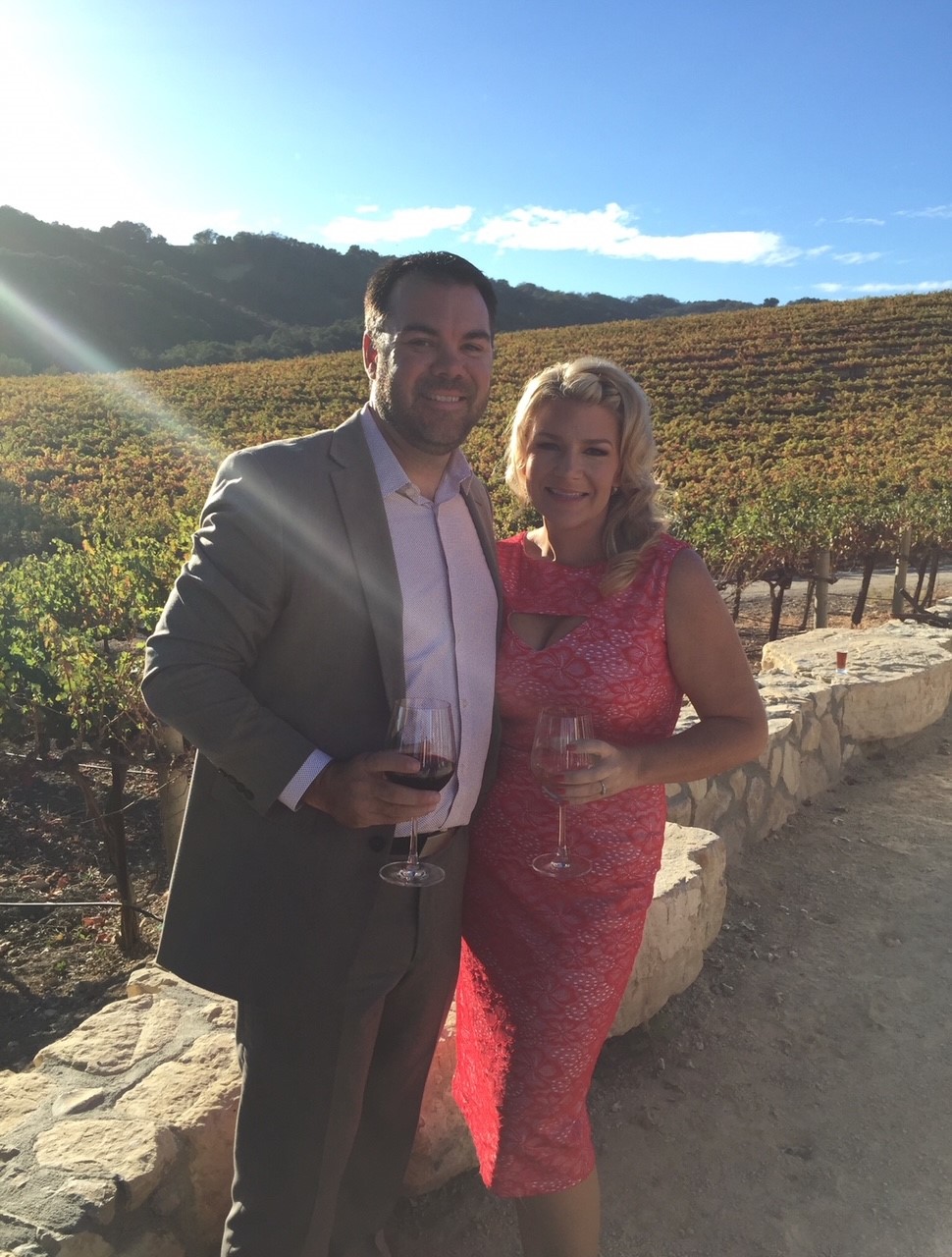 A man in a grey suit and a woman in a red dress smile while holding wine glasses in front of a sunlit vineyard. The background features rows of grapevines and hills under a clear blue sky.