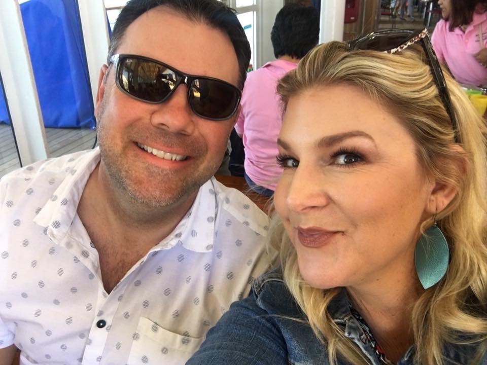 A man in sunglasses and a white patterned shirt smiles alongside a woman with blonde hair wearing blue earrings and a denim jacket. They are both outdoors, seated, with other people in the background.