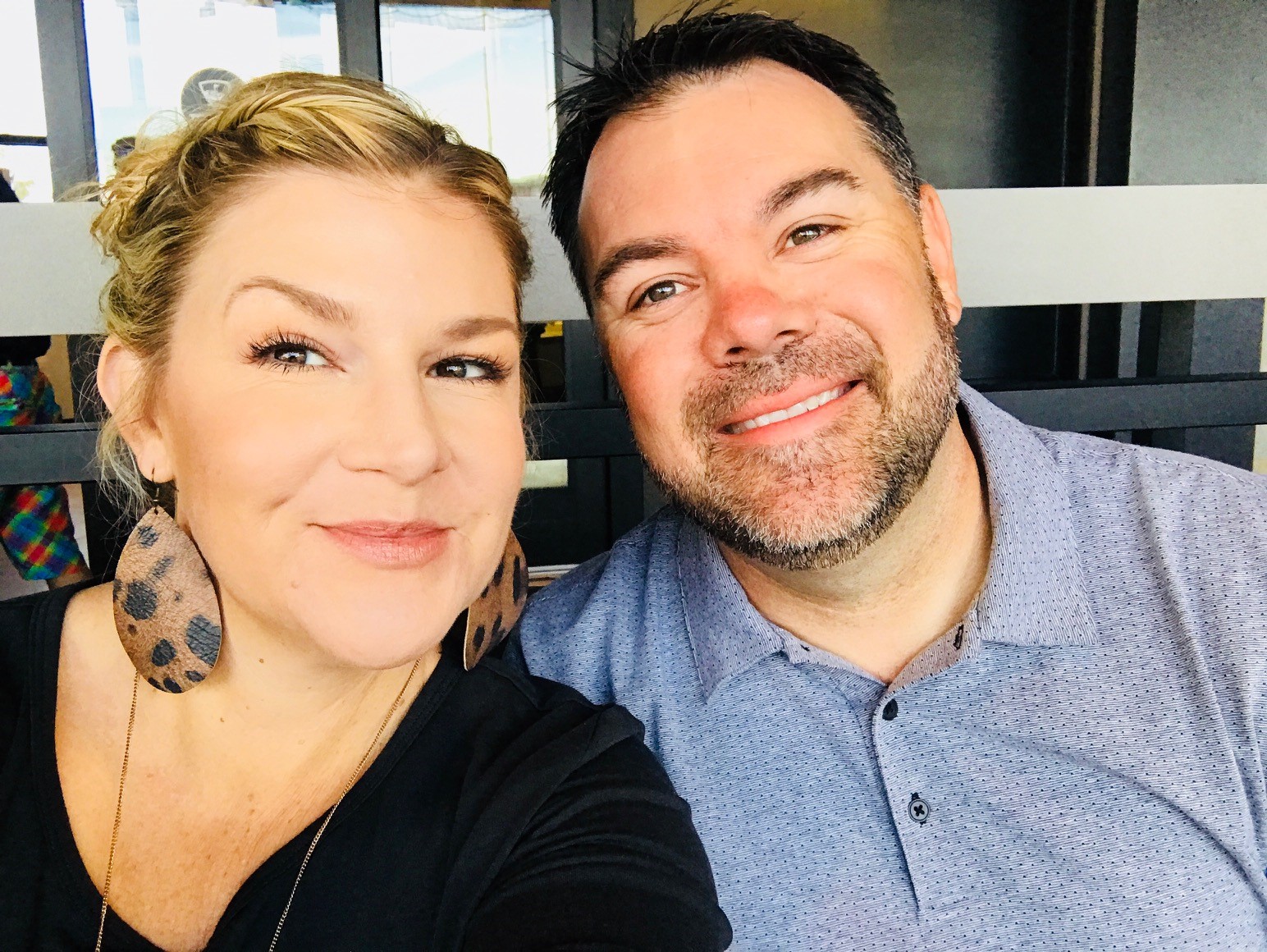 A man and woman are smiling at the camera. The woman is wearing large, dangling earrings with a spotted pattern and has her hair styled. The man has a short beard and is wearing a light-colored polo shirt. They appear to be sitting indoors.