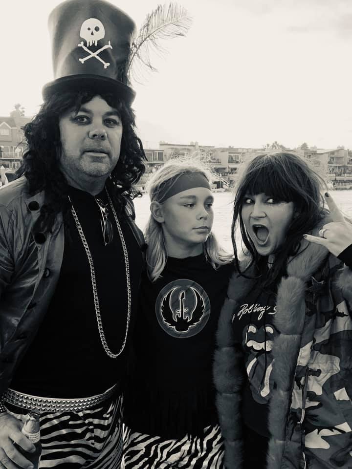 A black-and-white photo shows three people in costume. The individual on the left wears a tall hat decorated with a skull and crossbones, and the person in the middle has long hair and a headband. The person on the right, with a wig, is making a playful open-mouthed expression.