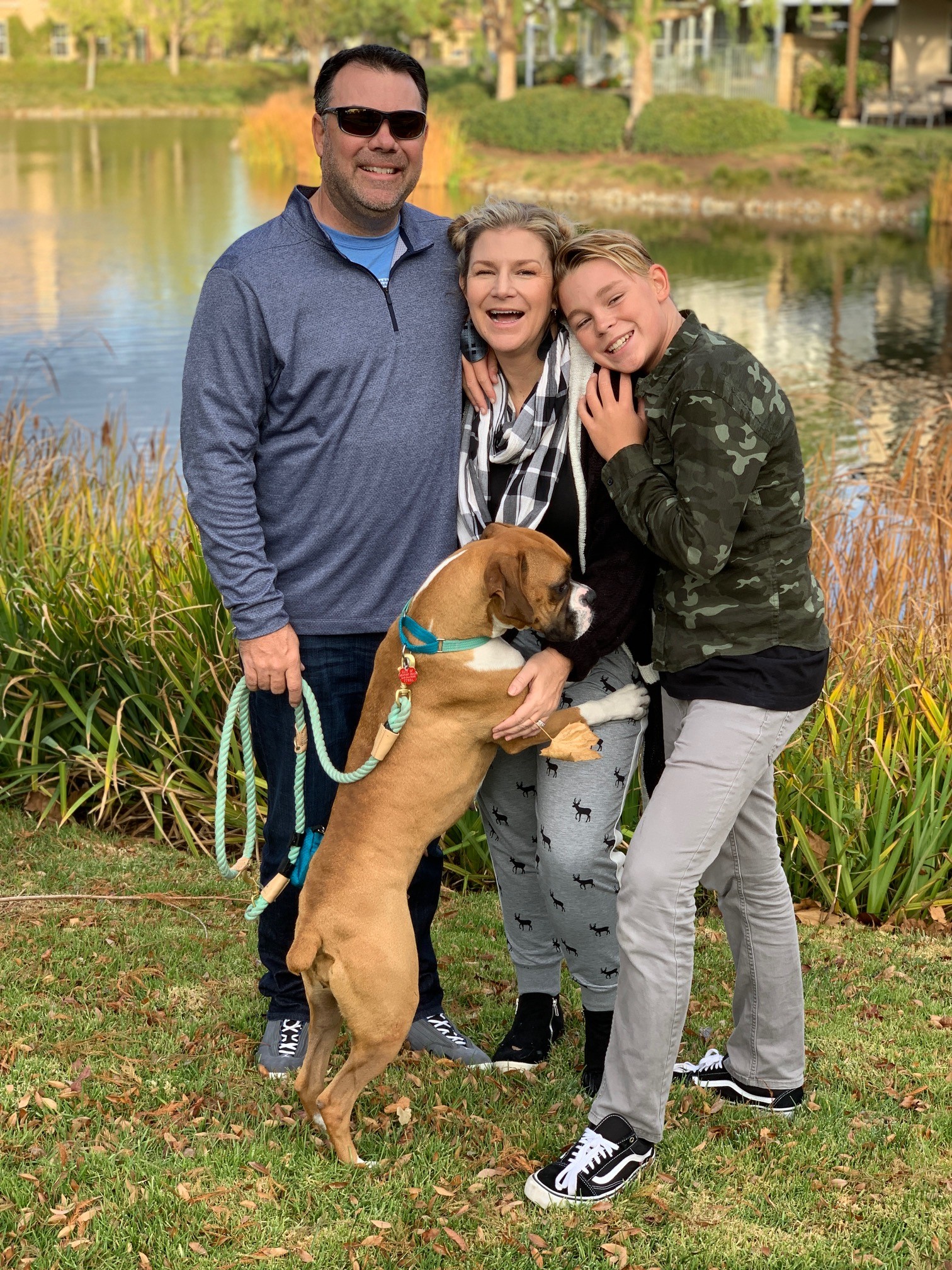 A smiling family of three poses outdoors by a pond. The father is holding a large brown dog on a leash. The mother and child are hugging and laughing. The mother is wearing a scarf and the child is dressed in a camouflage jacket. Green grass and reeds can be seen.