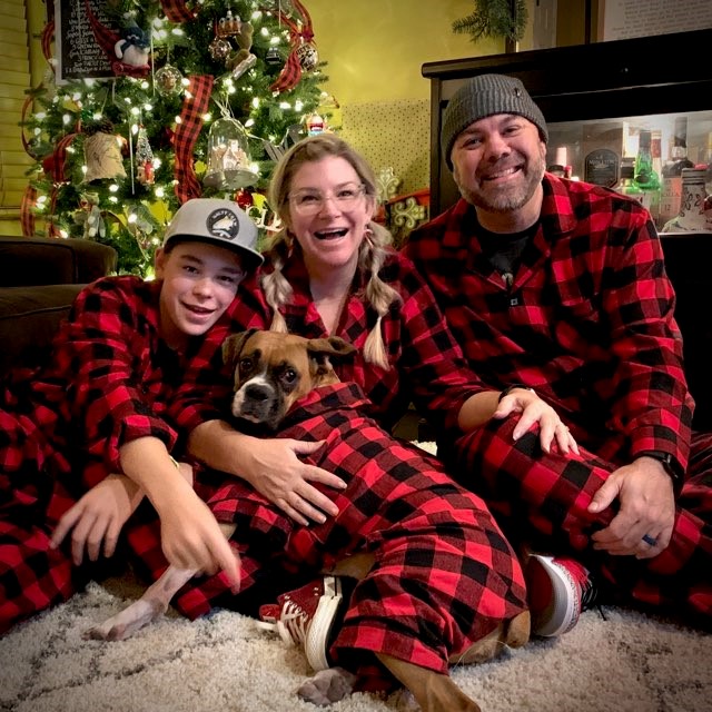 A family of three and their dog sit on the floor in front of a Christmas tree, all wearing matching red and black plaid pajamas. They are smiling and appear joyful, celebrating the holiday season together. The tree is decorated with lights and ornaments.