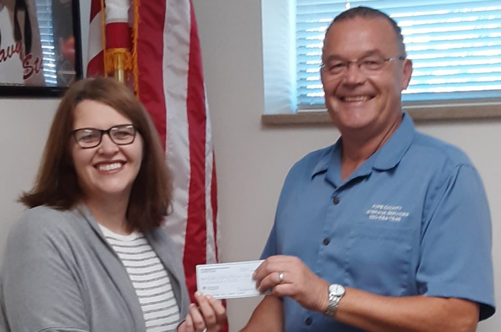 A woman with glasses and a man in a blue shirt are smiling while holding a check together. Behind them is an American flag and a window with blinds partially open.