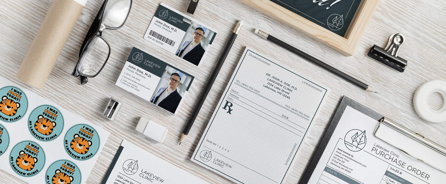 A neatly arranged home office desk contains glasses, ID cards, business cards, a clipboard with a purchase order, stickers with tiger faces, a roll of paper, a pencil, a pen, and an eraser. Branded items with Lakeview Clinic's logo are prominently displayed.