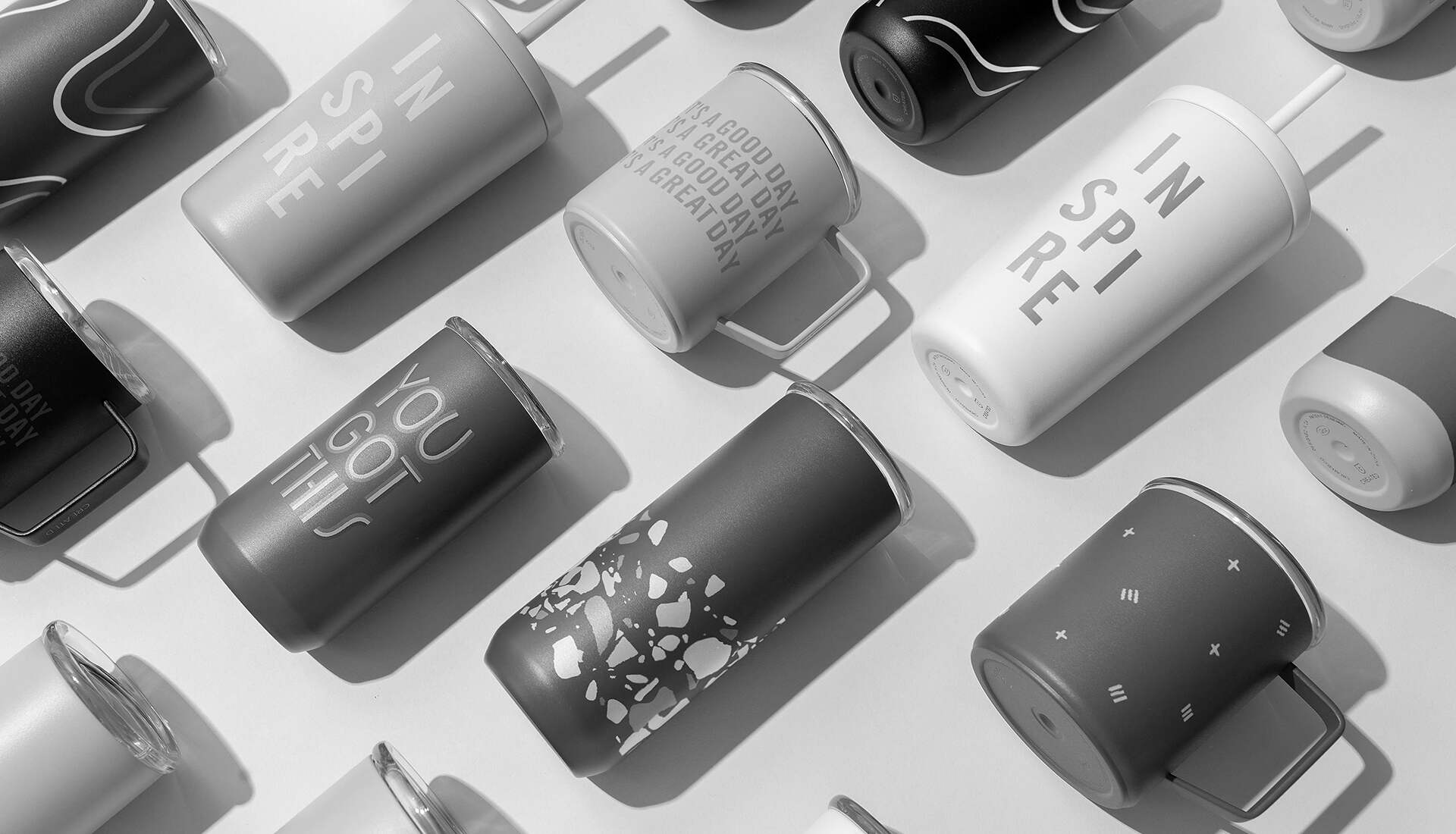 Top view of various reusable travel mugs and tumblers arranged on a light background. The mugs have different designs and motivational phrases like "INSPIRE" and "YOU GOT THIS" in a mix of monochrome colors and patterns.