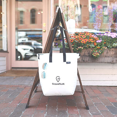 A white GroundTruth tote bag hangs from a wooden stand on a brick sidewalk in front of a storefront. The bag has a pair of sunglasses clipped to the front, and the shop entrance is decorated with vibrant flowers.