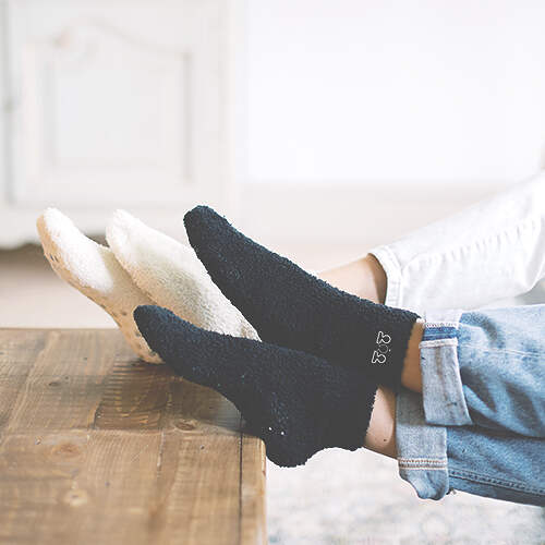 Two individuals are relaxing on a couch with their feet propped up on a wooden coffee table. They are wearing cozy, fluffy socks; one pair is black with a small design near the ankle, and the other pair is white. The lower part of their jeans is visible.