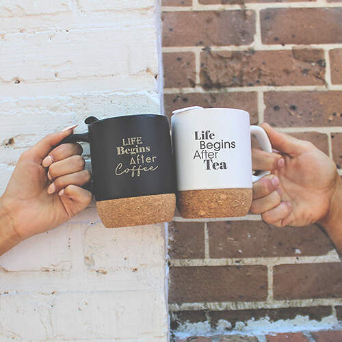 Two people clink mugs against a brick and white-painted wall, embodying a shared culture of morning rituals. The left black mug reads "Life Begins After Coffee," and the right white mug reads "Life Begins After Tea." Both mugs feature cork bases.