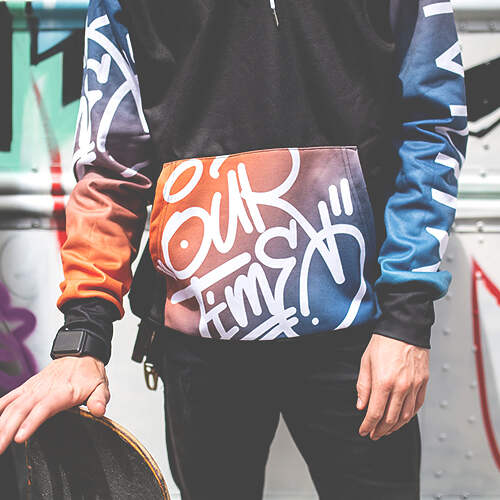 A person wearing a colorful hoodie with abstract graffiti designs stands in front of a graffiti-covered wall, holding a skateboard. The hoodie has a front pocket with vibrant orange, red, blue, and purple patterns and white, abstract lettering.