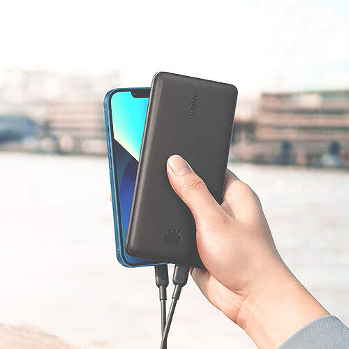 A hand holding a black Anker power bank and a smartphone, both connected by charging cables. The smartphone screen is partially visible and turned on. The background shows a blurred outdoor setting with what appears to be buildings and a body of water.
