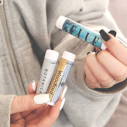 A person with black and white nail polish holds three lip balm tubes in their hands. The tubes are labeled "BOTANICA," "Woodford," and "FLIP." The person is wearing a light gray jacket. The background is blurred.