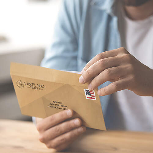 A person in a light blue shirt is opening a tan envelope. The envelope has a Lakeland Realty logo on the back flap and is addressed to John Doe, 2349 Main Street, Lakeville, CA, 51234. The envelope also features a U.S. flag stamp.