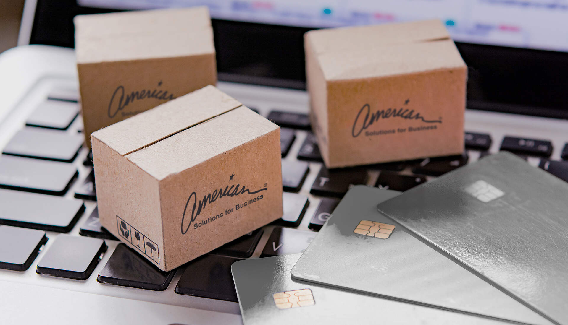 Four small cardboard boxes with "American Solutions for Business" written on them are placed on a laptop keyboard, embodying a culture of corporate efficiency. Three credit cards with visible chip technology lie beside the boxes on the keyboard.