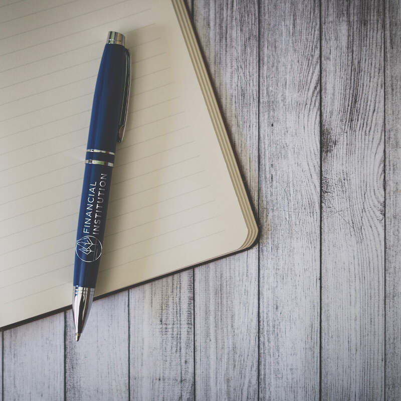 A blue pen with "Financial Institution" printed on it lies on the open pages of a lined notebook, which is set on a wooden surface with a rustic texture. The scene suggests a workspace or a moment of jotting down notes or ideas about markets.