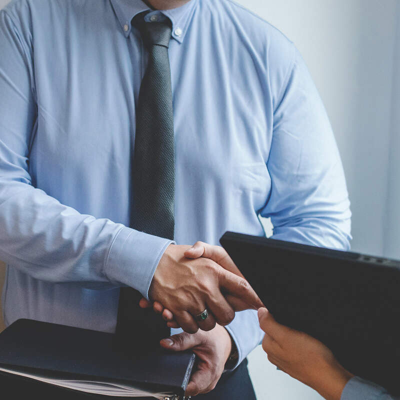 Two people shaking hands. One person is dressed in a light blue dress shirt and dark tie, holding a portfolio. The other individual, holding a tablet with market data displayed, has an outstretched hand as they shake hands. The background is neutral and blurred.