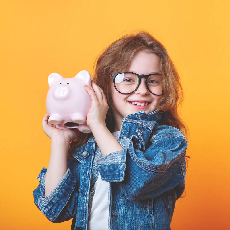 A young girl with long hair and glasses smiles while holding a pink piggy bank next to her face. She is wearing a denim jacket over a white shirt, ready to explore new markets. The background is a vibrant yellow.