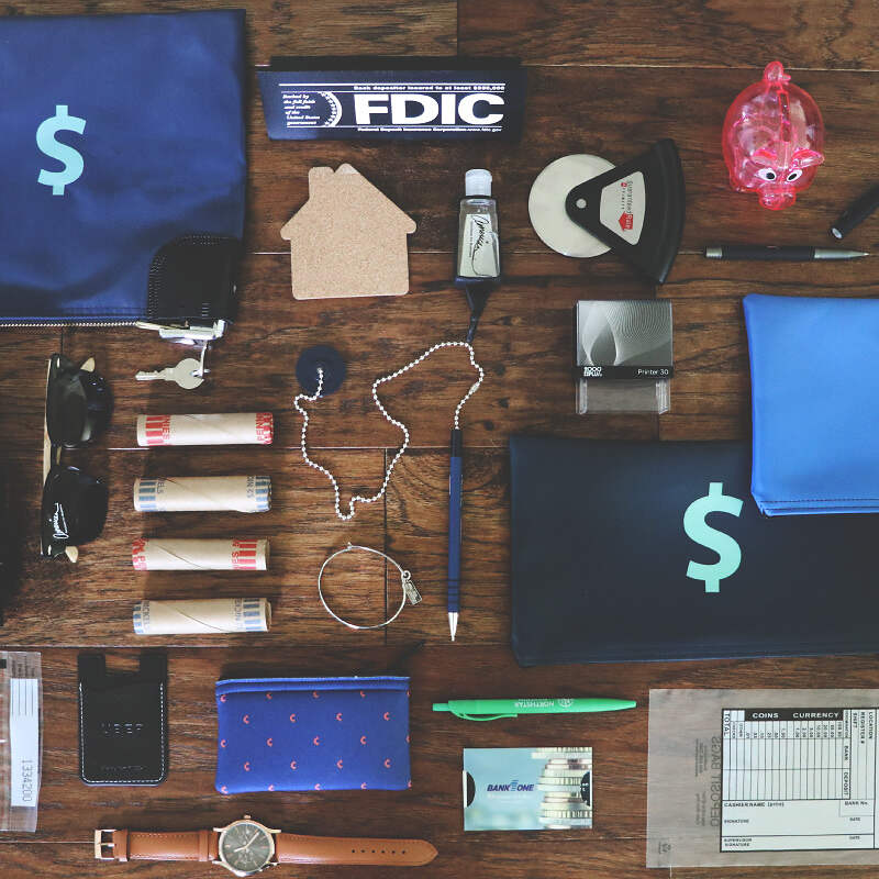 A flat lay of various financial and office items on a wooden surface. Items include blue pouches with dollar signs, an FDIC sign, a cork house shape, rolls of coins, a piggy bank, a wristwatch, a pen, sunglasses, and other small office supplies—all set to represent different aspects of markets and economy.