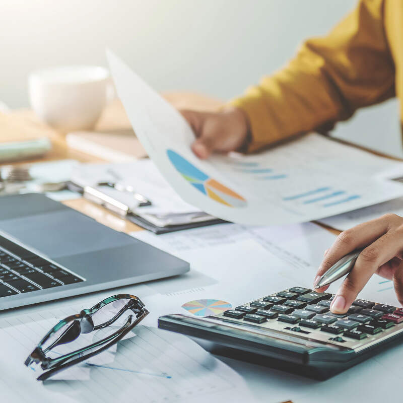 A person sits at a desk using a calculator with one hand and holding a piece of paper with a pie chart in the other. A laptop, glasses, and several documents are scattered on the desk, suggesting they are analyzing financial markets or planning investments.