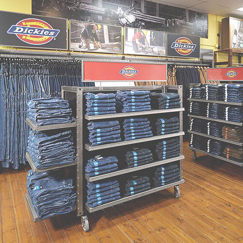 A clothing store display featuring shelves stocked with neatly folded blue jeans on a wooden floor. The shelves are organized with signage showing the Dickies brand, with additional hanging racks of jeans visible in the background.