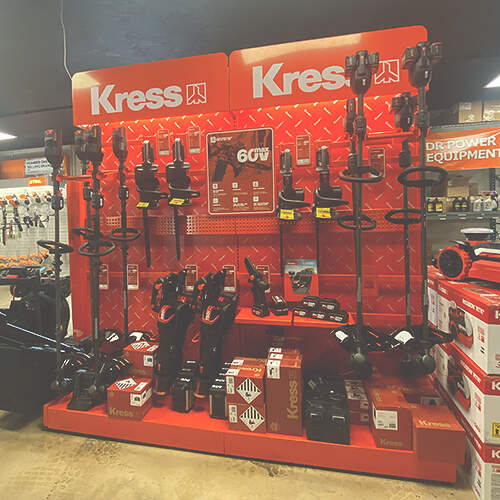 A store display featuring red and black Kress branded power tools, including several weed trimmers and other equipment, arranged on shelves and hooks against a bright red background. Signs with product information and prices are also visible.