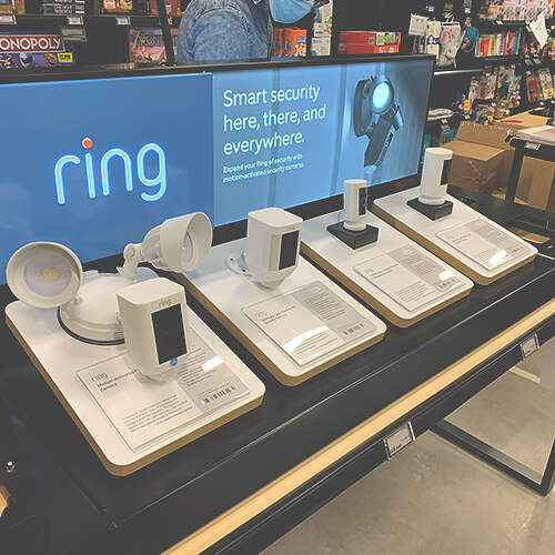 A display of various Ring smart security devices is shown in a store. The background features a screen with the Ring logo and the tagline, "Smart security here, there, and everywhere." Several models of cameras and lights are placed on individual stands.