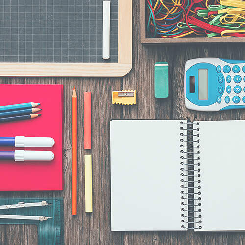A variety of school supplies arranged on a wooden surface, including a blank notebook, colored pencils, markers, a calculator, an eraser, a pencil sharpener, a chalkboard, a protractor, and a box of rubber bands.