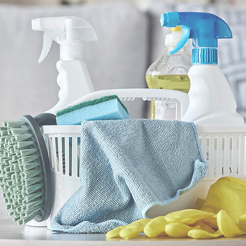 A cleaning supplies basket containing spray bottles, a scrub brush, a sponge, a microfiber cloth, a bottle of liquid cleaner, and a pair of yellow rubber gloves. The items are arranged neatly inside the white plastic basket.