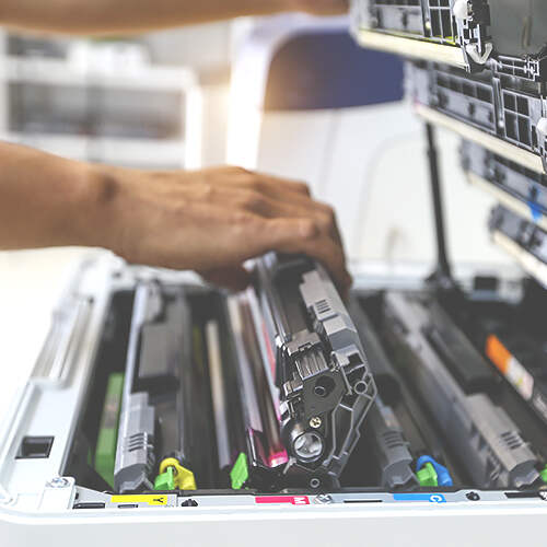 Close-up of a person’s hand inserting or removing a toner cartridge into/from a laser printer. Multiple toner cartridges in various colors are visible inside the printer's open compartment, indicating maintenance or replacement work.
