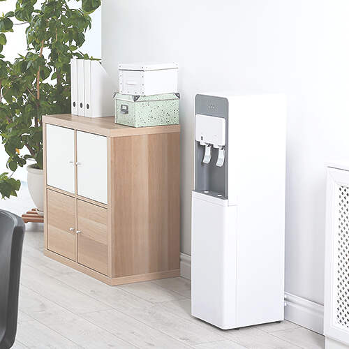 A modern office setup featuring a light wooden storage cabinet with white doors, a potted plant on top, and organized files and boxes. Next to it stands a white water dispenser against a light-colored wall. The floor is covered with light wood laminate.