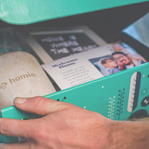A hand opening a teal box containing a wooden item with the text "homie," a card with images of people and text welcoming someone home, and another card with blurred text. The box has a nature-themed design with leaves and plants.