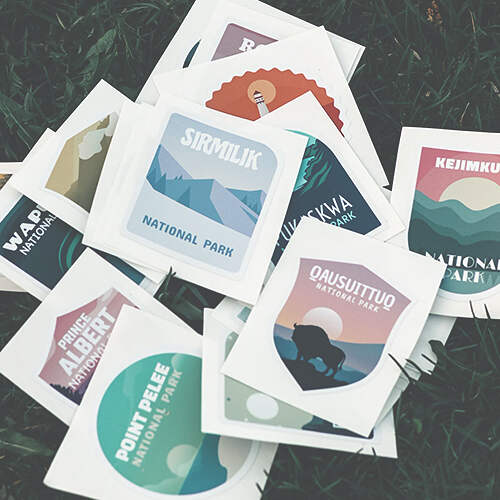 A pile of colorful national park stickers lies on the grass. The stickers depict various national parks with names such as Point Pelee, Sirilik, Kejimkujik, and Qausuittuq, among others, each with unique illustrations showcasing nature and landscapes.