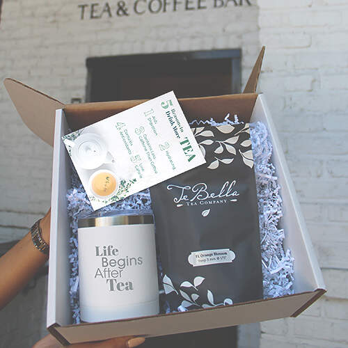 A person is holding an open gift box containing a white candle with the text "Life Begins After Tea," a packet of loose leaf tea from TeBella Tea Company, and an informational card about the benefits of tea. The box is filled with white crinkle paper.