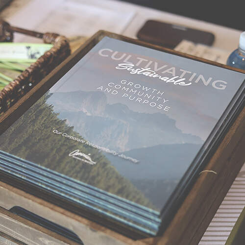 A stack of booklets is displayed on a wooden tray. The top booklet reads "Cultivating Sustainable Growth, Community, and Purpose" with an image of mountains in the background. Text at the bottom says "Our Corporate Responsibility and Journey.