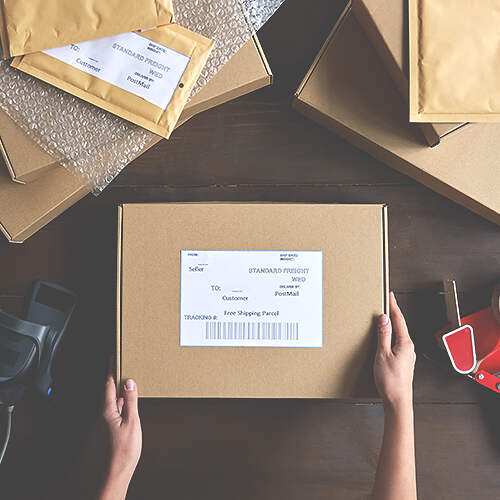 A pair of hands holding a closed cardboard box with a shipping label on a wooden surface. The table is surrounded by other shipping materials including bubble mailers, taped boxes, rolls of tape, a tape dispenser, and a black marker.