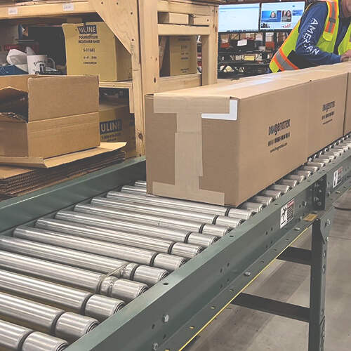 A long, rectangular cardboard box is moving along a conveyor belt in a warehouse-like setting. Nearby, shelving holds more cardboard boxes and various supplies. A person in a safety vest is visible in the background next to computer monitors.