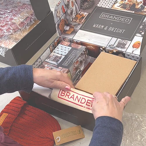 A person is assembling a branded gift box that includes a red knitted beanie, a cork notebook, a branded sticker, and some other branded items. The box features the words "BRANDED" and "WARM & BRIGHT" on the inside flaps. Bubble wrap is present on the side.