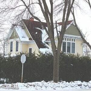 A two-story yellow house with white trim, partially obscured by trees and a hedge, is covered in snow. The roof has some snow patches, and a stop sign is visible in the foreground. This cozy home sits in a snowy, winter landscape that captures the serene essence of winter culture.