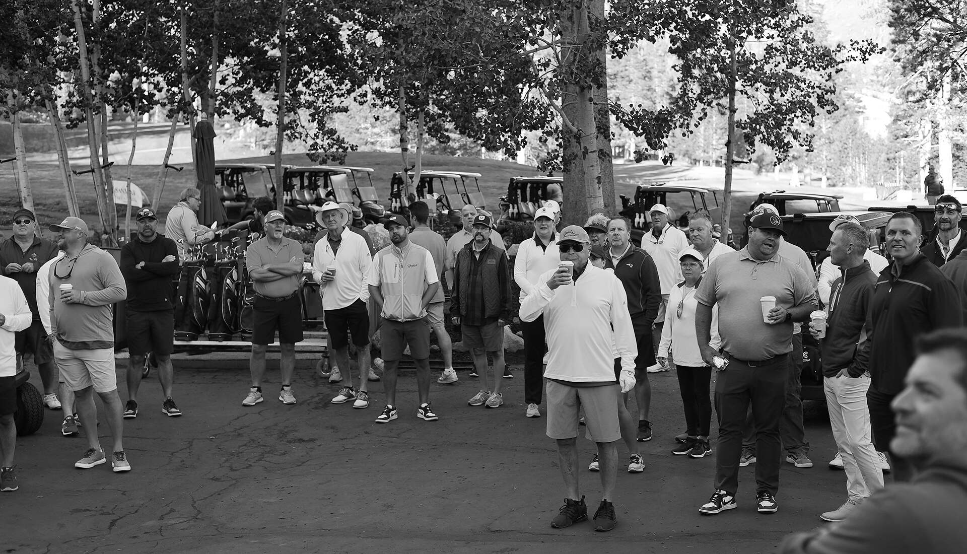 A group of people gather outdoors, among trees and golf carts, many holding beverages. Most are wearing casual attire, including caps and sneakers. The setting appears to be a golf course parking area. The mood is relaxed and social, reflecting a casual culture. The image is in black and white.