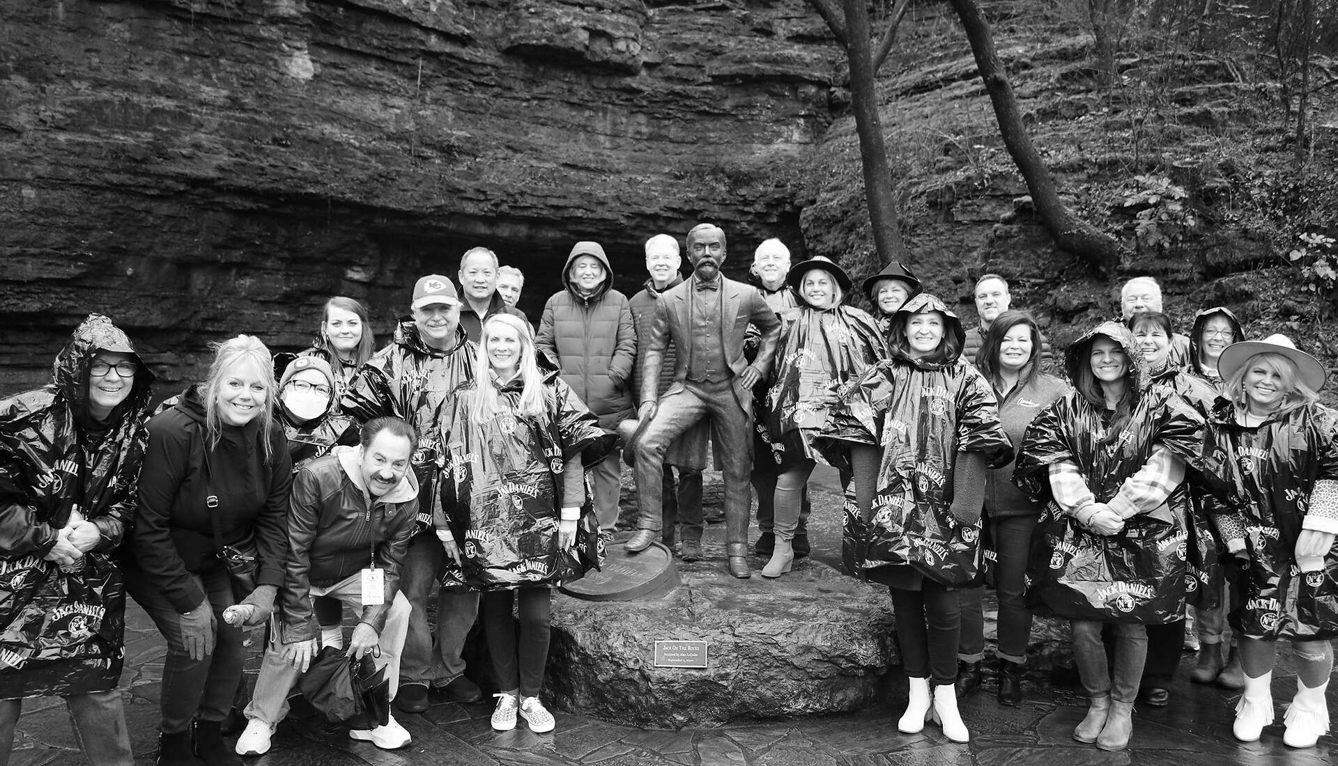 A group of people stands in front of a statue of a man in an outdoor setting, likely a park. Many are wearing rain ponchos, indicating wet weather. The statue is perched on a rock base, and the background features a rocky, natural landscape with trees. It's a moment immersed in both nature and culture.