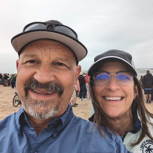 A smiling man and woman, Mike Schmitz and his friend, wearing hats and glasses take a selfie on a beach. The man has a mustache and the woman has long hair. They are dressed in casual clothes, with other people in the background under overcast skies.