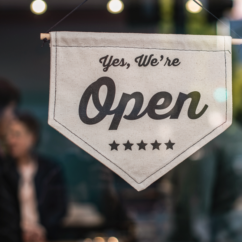 A white, triangular banner with the text "Yes, We're Open" and five stars hangs in a market window, indicating that the store is open for business. The background shows blurred silhouettes of people and a softly lit interior.