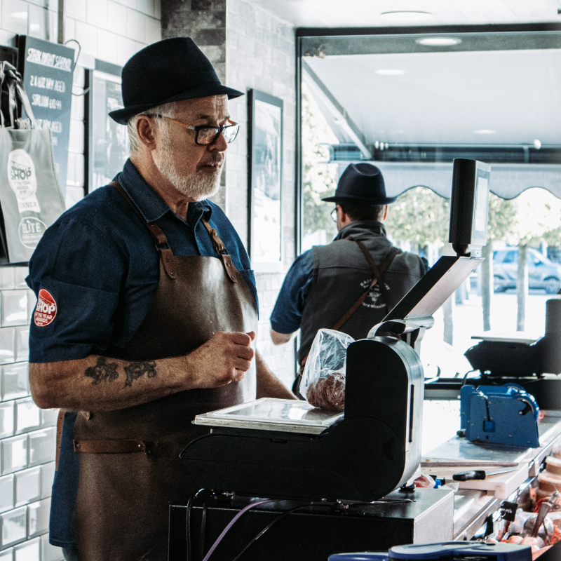 A man wearing a hat and apron stands behind a cash register at a counter in the market, holding a small bag. Another person is visible in the background, also wearing a hat and apron. The shop has a window through which outdoor foliage can be seen.